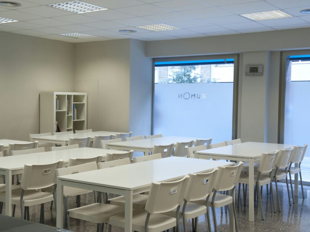 Aula Moderna con Abundante Luz Natural Una aula espaciosa y bien iluminada con mesas blancas dispuestas en filas, rodeadas de sillas de plástico blancas. Las paredes están pintadas en un tono neutro claro, y una gran ventana llena la habitación de luz natural. En el fondo se puede ver una estantería simple llena de libros, destacando un entorno de aprendizaje limpio y organizado.