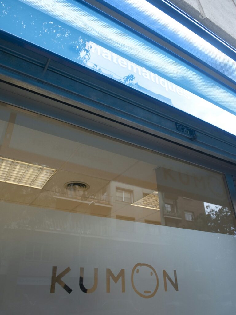 Entrada del Centro Educativo Kumon en Barcelona - Diseño Moderno y Funcional Fachada del Centro Kumon en Barcelona, con diseño contemporáneo y logo dorado en la puerta de cristal. Reflejos de la arquitectura circundante y luz natural iluminan el espacio.
