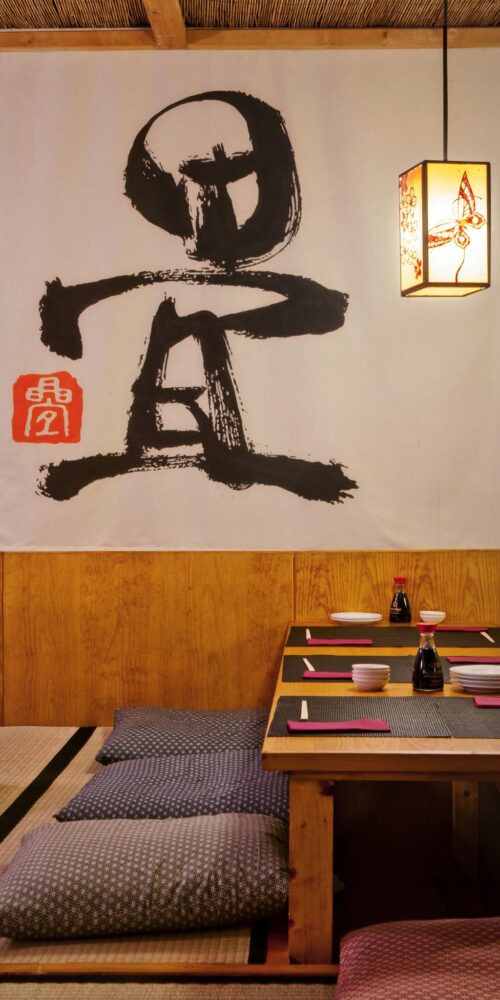 Área de comedor en estilo japonés con mesa baja de madera, platos y palillos, rodeada de tatamis y cojines, iluminada por una lámpara de diseño delicado y con un toque artístico gracias a un colgante caligráfico en la pared.