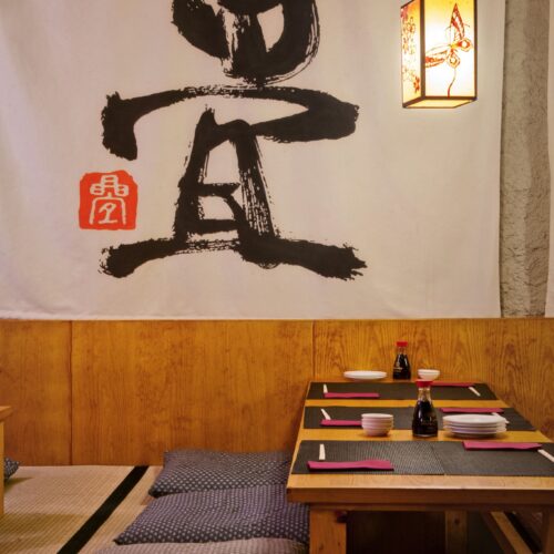 Área de comedor en estilo japonés con mesa baja de madera, platos y palillos, rodeada de tatamis y cojines, iluminada por una lámpara de diseño delicado y con un toque artístico gracias a un colgante caligráfico en la pared.