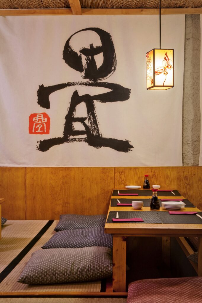 Área de comedor en estilo japonés con mesa baja de madera, platos y palillos, rodeada de tatamis y cojines, iluminada por una lámpara de diseño delicado y con un toque artístico gracias a un colgante caligráfico en la pared.