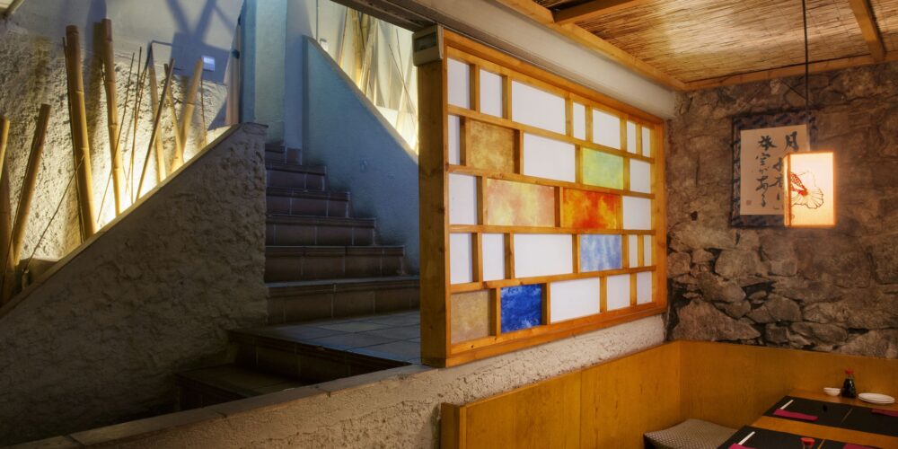 Escalera iluminada que conduce a un área de comedor acogedora en un restaurante de Barcelona, combinando elementos de diseño moderno y tradicional, con paneles de vidrio de colores y mobiliario de madera.