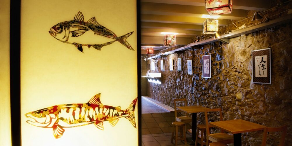 Comedor contemporáneo en Barcelona con decoración única, incluyendo farolillos de papel iluminados y motivos artísticos de peces en una partición. La pared de piedra rústica complementa la atmósfera acogedora del espacio, que cuenta con mesas y sillas de madera dispuestas para cenar, reflejando la funcionalidad y la estética moderna.