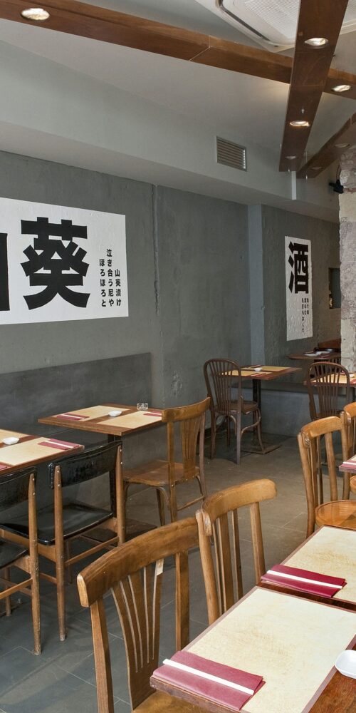 Interior de un restaurante moderno en Barcelona con mesas y sillas de madera, iluminación natural y un ambiente industrial. Las paredes presentan texto en caracteres asiáticos y los mesas están decoradas con manteles y pequeños cuencos.