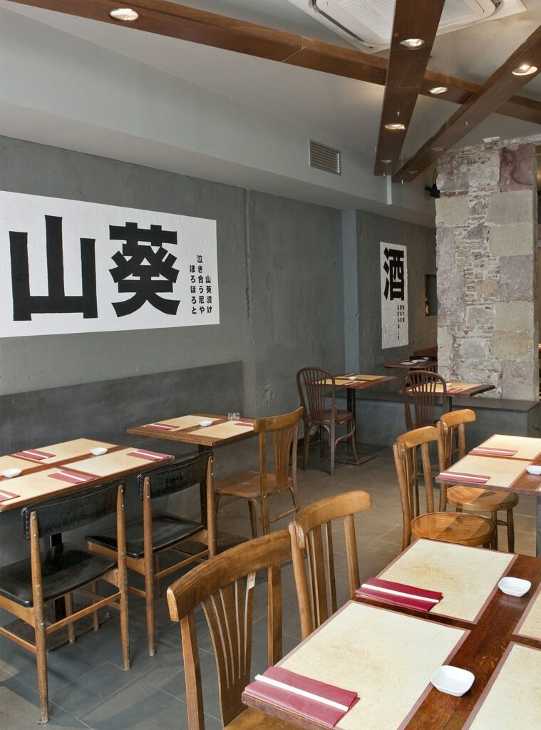 Diseño Moderno de Restaurante en Barcelona con Estilo Minimalista Interior de un restaurante moderno en Barcelona con mesas y sillas de madera, iluminación natural y un ambiente industrial. Las paredes presentan texto en caracteres asiáticos y los mesas están decoradas con manteles y pequeños cuencos.