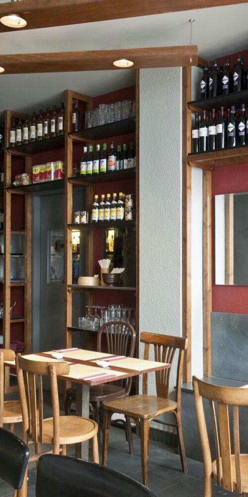 Diseño interior de un restaurante moderno en Barcelona con estanterías de madera llenas de botellas y copas, mesas rústicas de madera con cubiertos, y una estética contemporánea que resalta la iluminación natural y detalles arquitectónicos sofisticados.