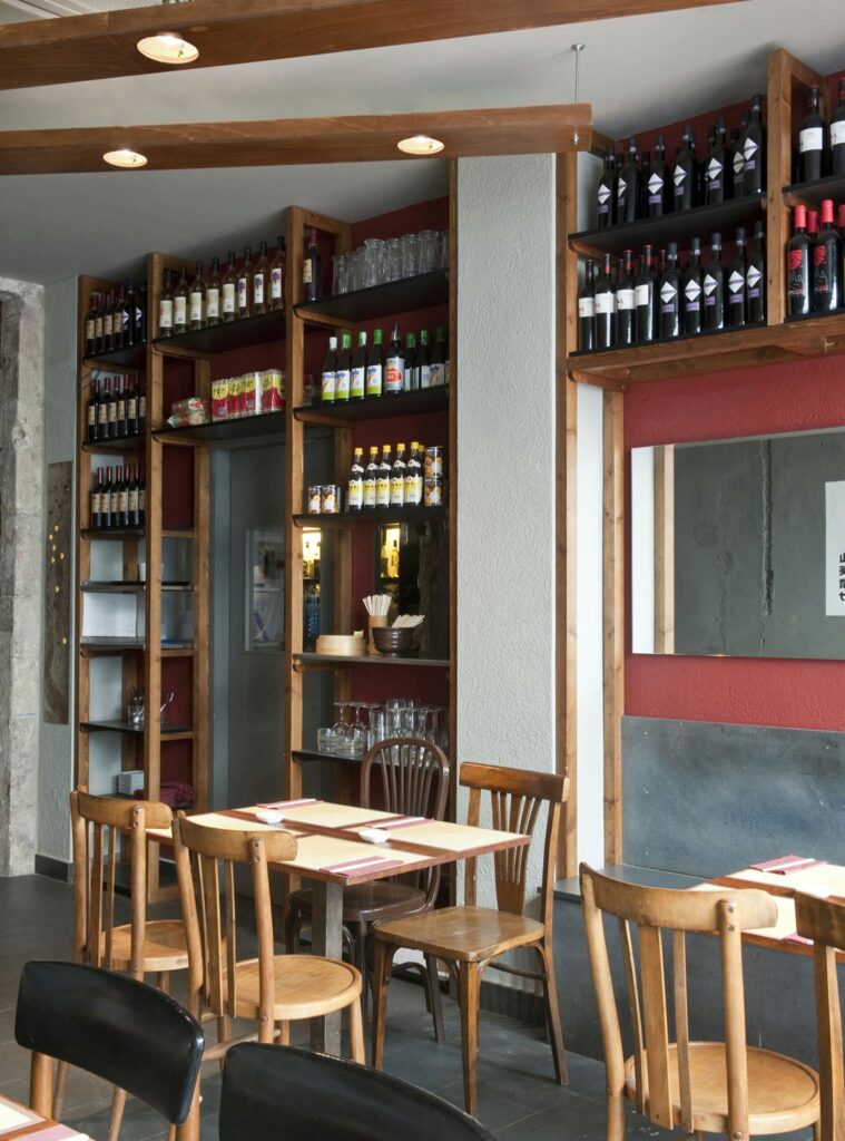 Diseño de interiores moderno en un restaurante de Barcelona Diseño interior de un restaurante moderno en Barcelona con estanterías de madera llenas de botellas y copas, mesas rústicas de madera con cubiertos, y una estética contemporánea que resalta la iluminación natural y detalles arquitectónicos sofisticados.