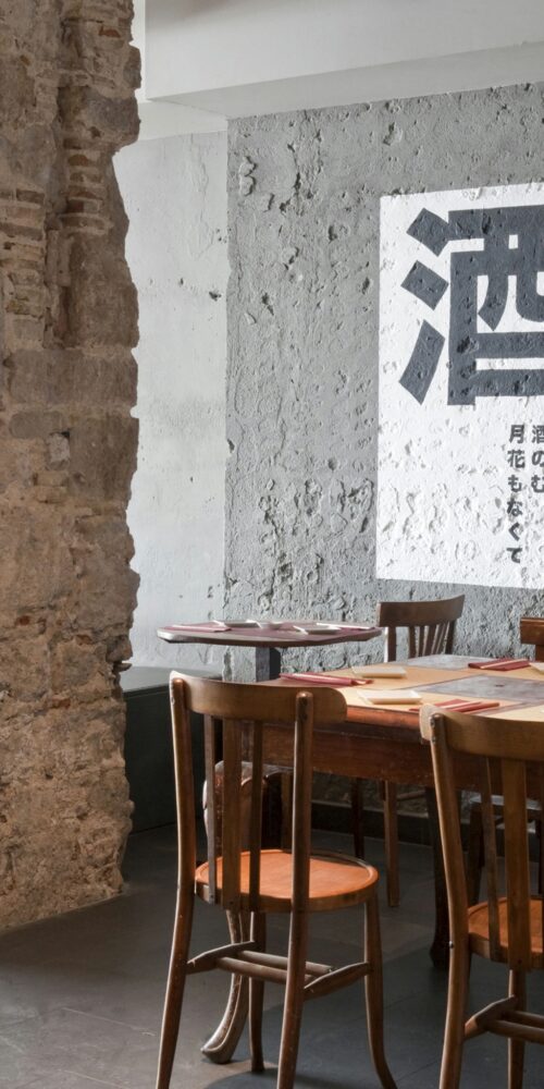 Comedor moderno en Barcelona con paredes de piedra rústica y una mesa de madera elegante con sillas, decorado con una obra de arte en blanco y negro. El espacio destaca la luz natural y detalles arquitectónicos sofisticados, fusionando funcionalidad y estética.