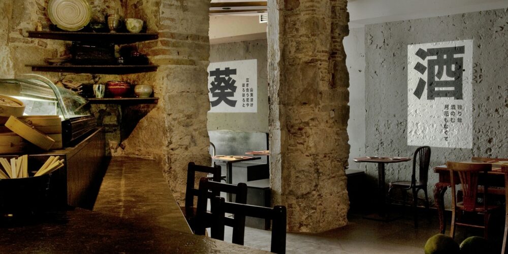 Interiores de un restaurante en Barcelona con diseño moderno, barra de madera oscura, utensilios decorativos, paredes de ladrillo expuesto, cemento y mesas de estilo clásico. Elementos gráficos asiáticos en las paredes y suave iluminación que resalta la estética elegante.