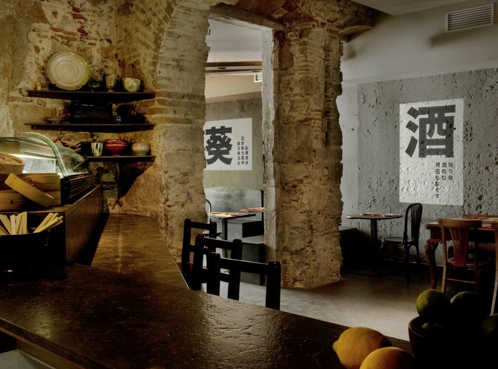 Diseño moderno y contemporáneo en restaurante de Barcelona Interiores de un restaurante en Barcelona con diseño moderno, barra de madera oscura, utensilios decorativos, paredes de ladrillo expuesto, cemento y mesas de estilo clásico. Elementos gráficos asiáticos en las paredes y suave iluminación que resalta la estética elegante.