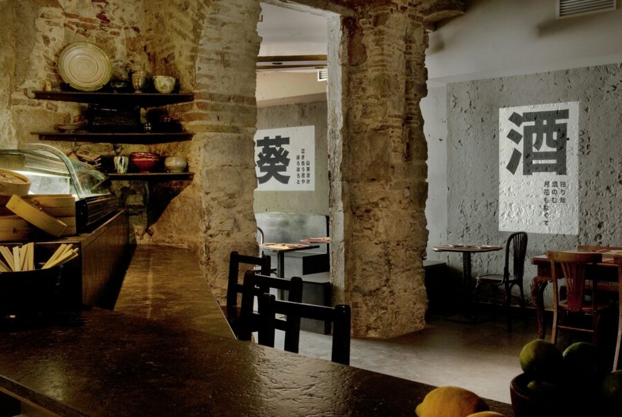 Interiores de un restaurante en Barcelona con diseño moderno, barra de madera oscura, utensilios decorativos, paredes de ladrillo expuesto, cemento y mesas de estilo clásico. Elementos gráficos asiáticos en las paredes y suave iluminación que resalta la estética elegante.