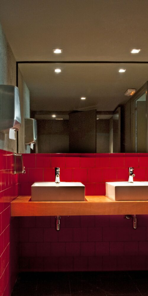Interior moderno de un baño en Barcelona con dos lavabos rectangulares sobre una encimera de madera, azulejos rojos brillantes, accesorios de acero inoxidable y iluminación empotrada.
