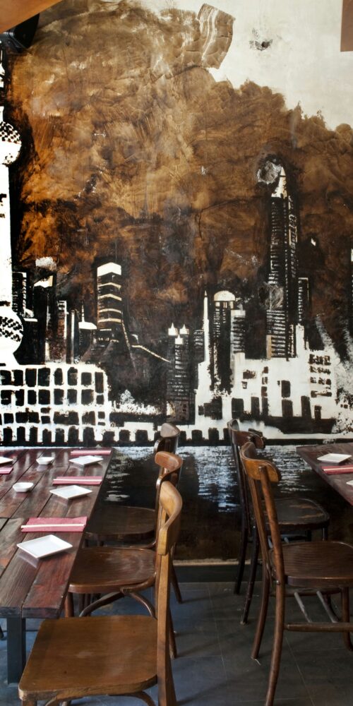 Diseño contemporáneo de un restaurante con mesas y sillas de madera rústica, mural monocromático de skyline urbano y mesas decoradas con platos blancos y servilletas rojas.
