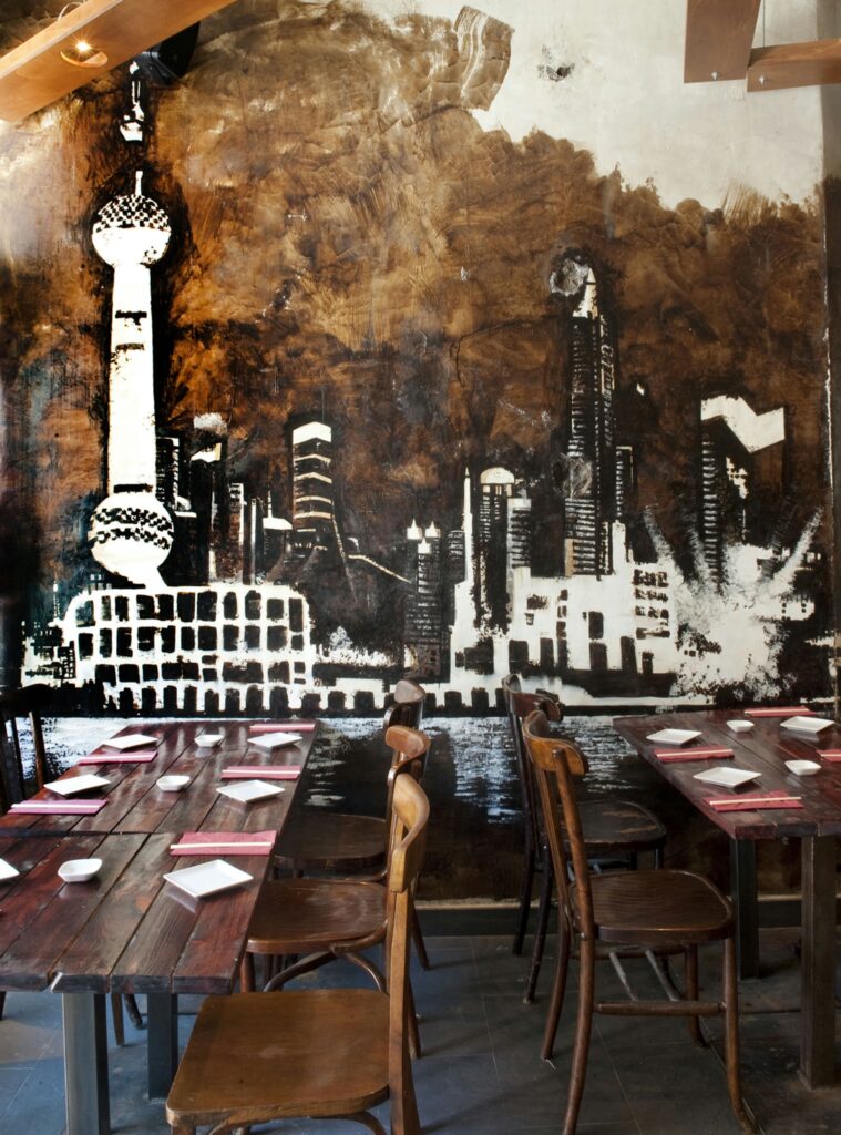 Diseño Interior de Restaurante Moderno en Barcelona Diseño contemporáneo de un restaurante con mesas y sillas de madera rústica, mural monocromático de skyline urbano y mesas decoradas con platos blancos y servilletas rojas.