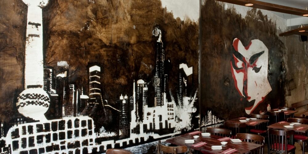 Vista interior de un comedor moderno en Barcelona con murales de una skyline y una cara estilizada, mesas de madera y sillas rojas, creando un ambiente acogedor y contemporáneo.