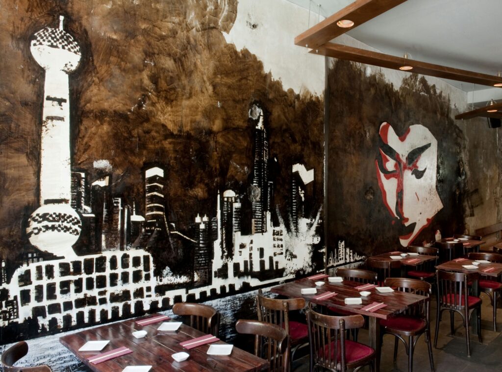 Comedor moderno con murales artísticos en Barcelona Vista interior de un comedor moderno en Barcelona con murales de una skyline y una cara estilizada, mesas de madera y sillas rojas, creando un ambiente acogedor y contemporáneo.