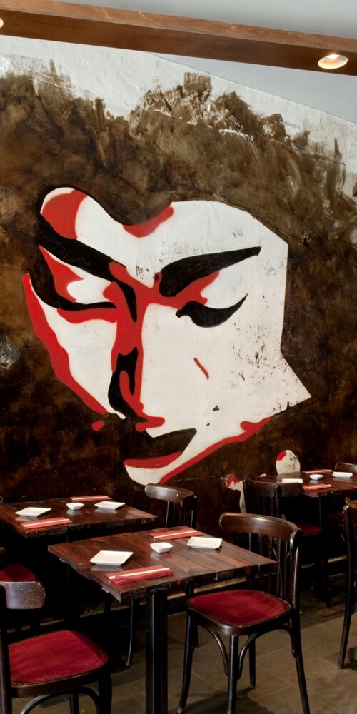 Interiores de un restaurante contemporáneo en Barcelona, destacando un mural grande de un rostro en tonos rojos y blancos, mesas de madera oscura y sillas tapizadas en rojo. Un espacio que combina funcionalidad y estética.