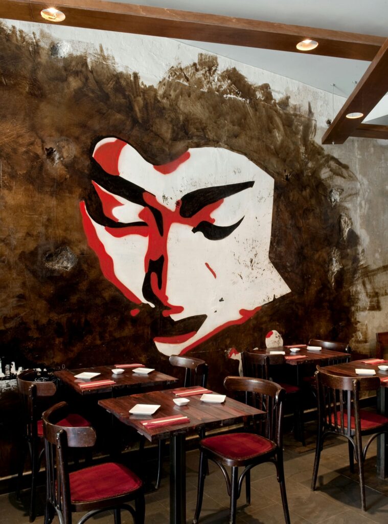 Diseño de Interiores de Restaurante Contemporáneo en Barcelona con Mural Artístico Interiores de un restaurante contemporáneo en Barcelona, destacando un mural grande de un rostro en tonos rojos y blancos, mesas de madera oscura y sillas tapizadas en rojo. Un espacio que combina funcionalidad y estética.