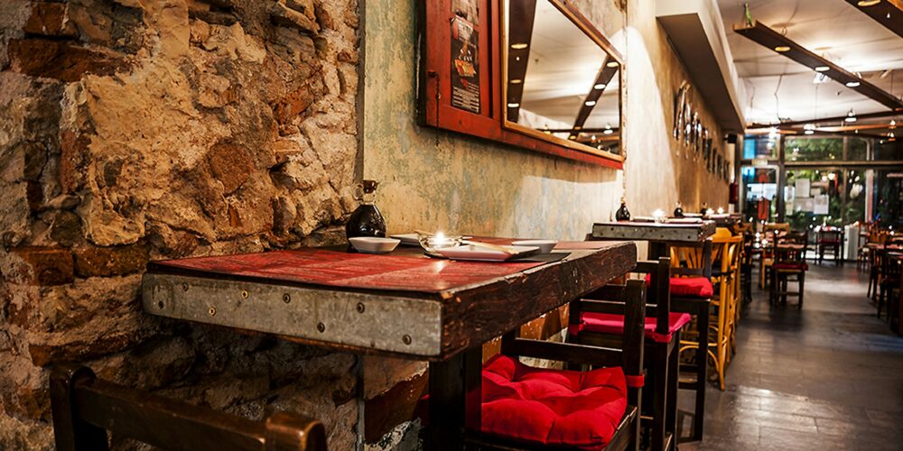 Vista interior de un restaurante contemporáneo en Barcelona, con paredes de piedra expuesta, mobiliario de madera y una mesa metálica con sillas rojas, destacando la funcionalidad y estética moderna.