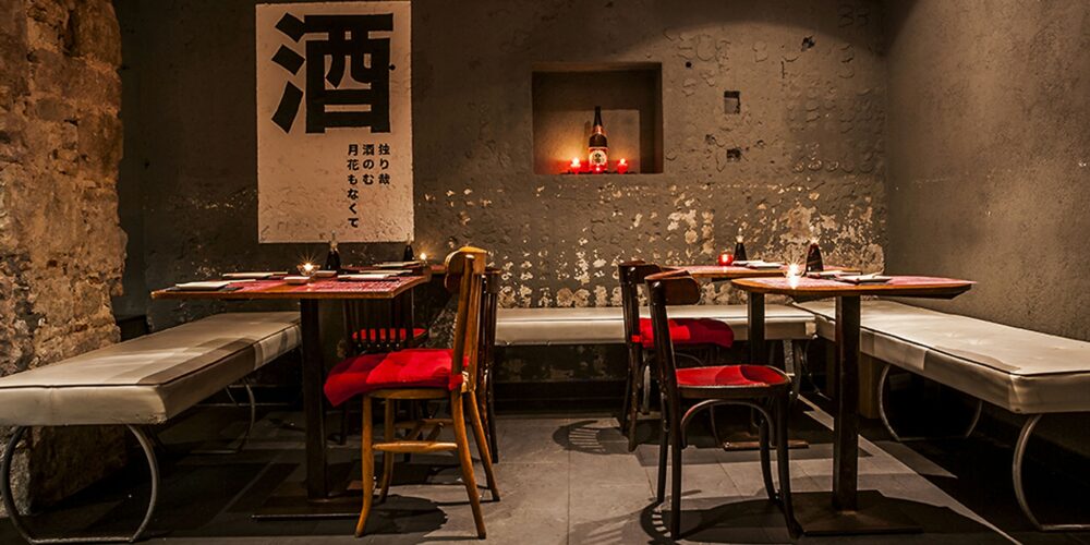 Interior de un restaurante moderno en Barcelona, con diseño elegante que incluye mesas de madera minimalistas, asientos con cojines rojos y blancos, y una pared de concreto decorada con arte de caligrafía japonesa. La iluminación ambiental resalta la funcionalidad y estética del espacio, creando un ambiente cálido y acogedor.