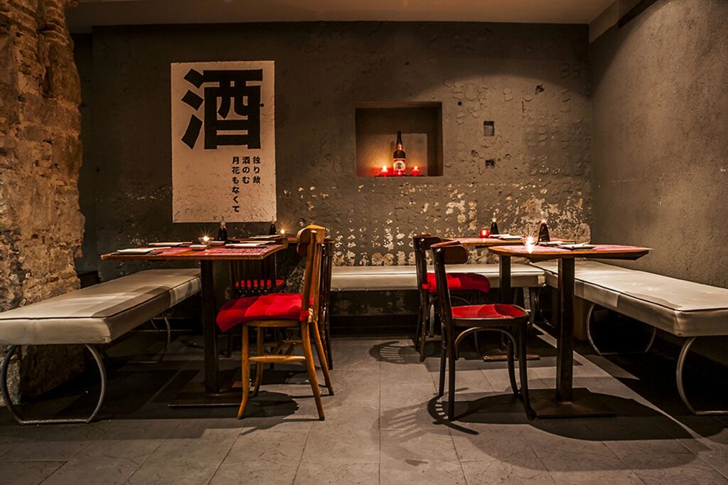 Diseño Interior Moderno de Restaurante en Barcelona Interior de un restaurante moderno en Barcelona, con diseño elegante que incluye mesas de madera minimalistas, asientos con cojines rojos y blancos, y una pared de concreto decorada con arte de caligrafía japonesa. La iluminación ambiental resalta la funcionalidad y estética del espacio, creando un ambiente cálido y acogedor.