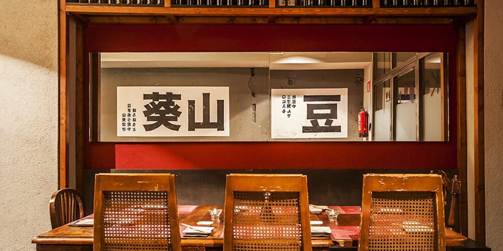 Diseño interior moderno de un restaurante en Barcelona, con una mesa de madera rústica, sillas de respaldo tejido y estantes que exhiben botellas de vino. La iluminación natural resalta detalles arquitectónicos y arte decorativo con caracteres japoneses, ideal para aficionados al diseño de interiores.
