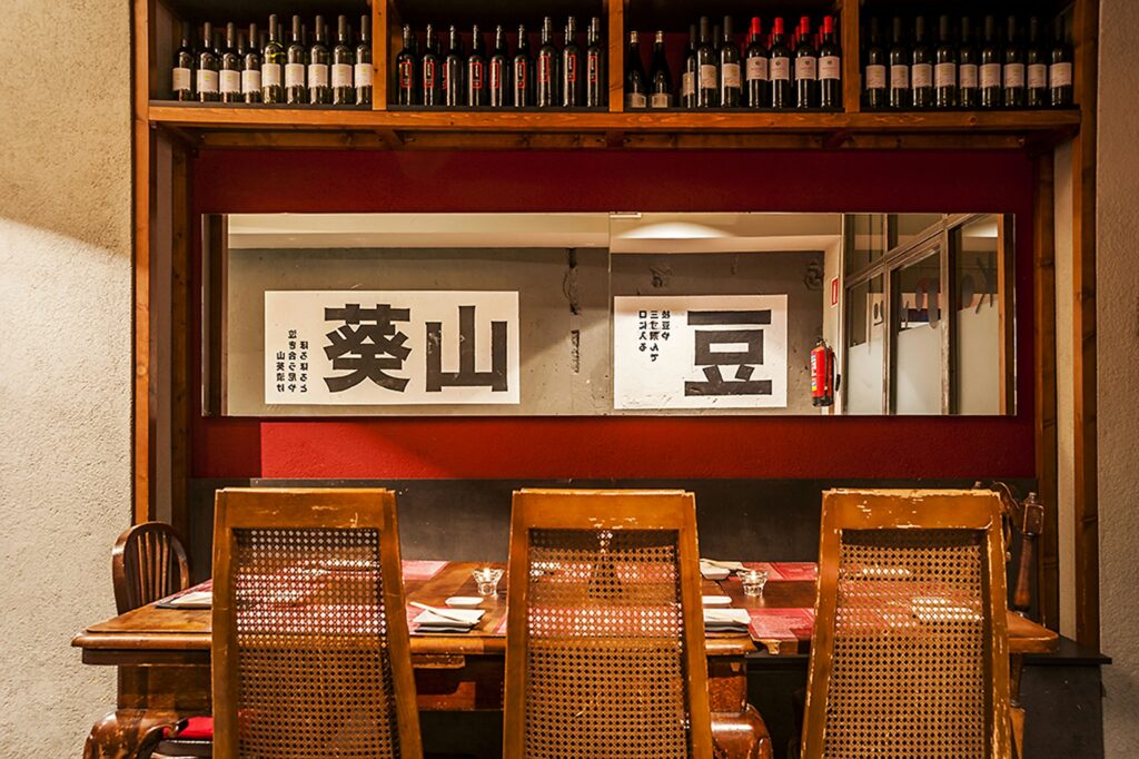 Interiores Modernos en un Restaurante de Barcelona con Toques Rústicos Diseño interior moderno de un restaurante en Barcelona, con una mesa de madera rústica, sillas de respaldo tejido y estantes que exhiben botellas de vino. La iluminación natural resalta detalles arquitectónicos y arte decorativo con caracteres japoneses, ideal para aficionados al diseño de interiores.