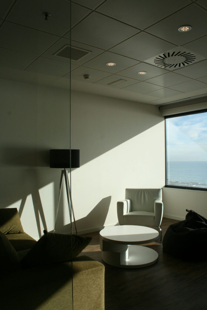 Espacio de Oficina Moderno y Acogedor con Ventana Grande y Luz Natural Un espacio de oficina moderno que cuenta con una gran ventana que permite que la luz natural ilumine la habitación. La luz del sol crea una sombra diagonal en el área, resaltando una mesa circular blanca posicionada en el centro. Cerca hay una silla de brazo de color gris claro y varios puff oscuros, proporcionando una opción de asientos casual. Una lámpara de pie elegante está colocada contra la pared, realzando la atmósfera acogedora del lugar de trabajo. El techo está diseñado con luces empotradas y ventilación, enfatizando la decoración minimalista que contribuye a un ambiente relajado perfecto para la productividad.