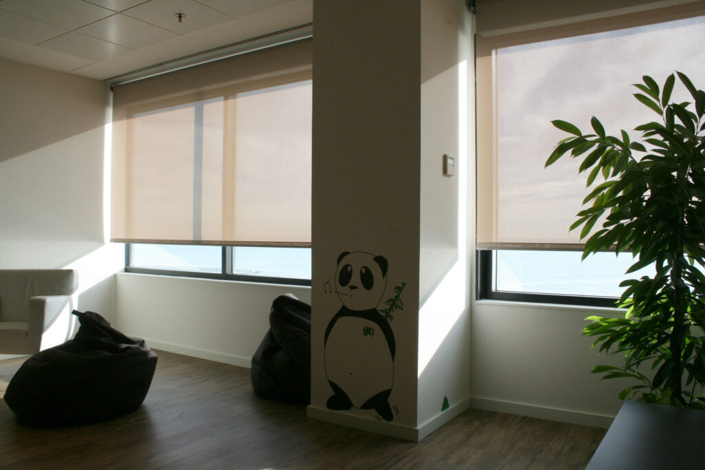 Interiores Modernos Acogedores con Arte de Panda y Luz Natural Una habitación moderna y luminosa con grandes ventanas adornadas con persianas enrollables de color beige claro. Una pequeña planta verde se encuentra cerca de la ventana derecha, mientras que una linda ilustración de panda decora la pared cercana. El suelo de madera clara añade calidez al espacio, complementado por cómodos sillones de frijol negros y un sofá gris simple, creando un ambiente acogedor perfecto para la relajación o como área de oficina.