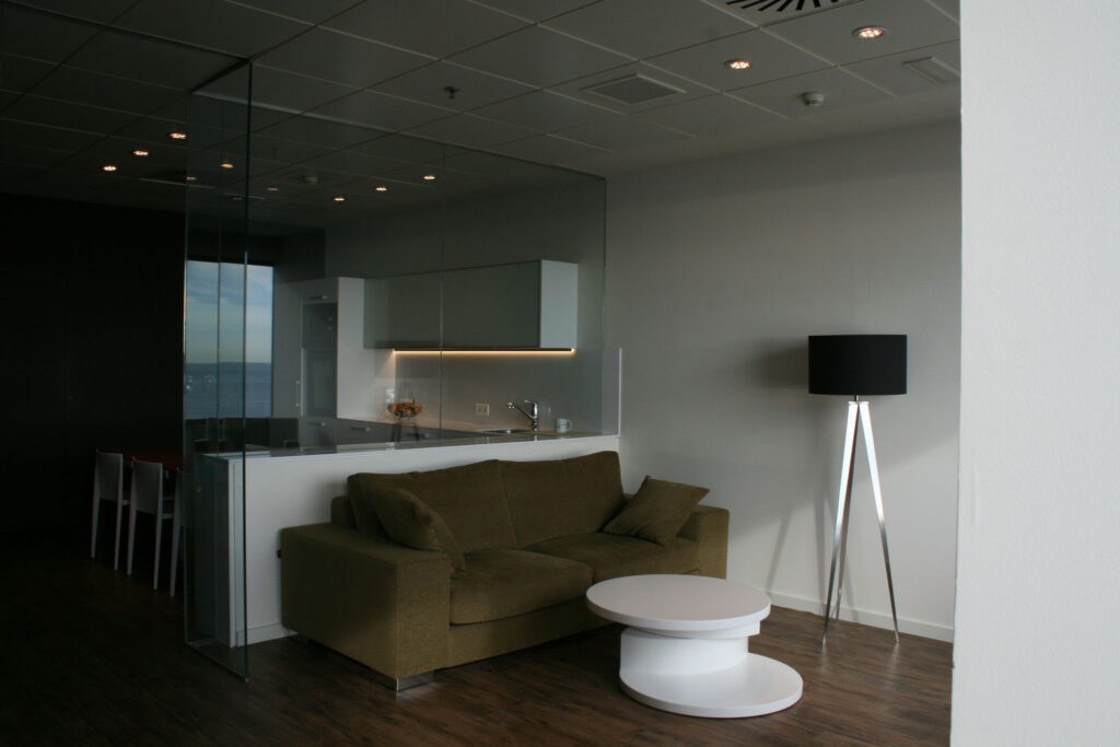 Espacio de Vida Moderno y Elegante con Sofá Verde y Diseño Minimalista Una sala de estar elegante que presenta un vibrante sofá verde con cojines decorativos, complementado por una mesa de café redonda blanca y minimalista. El fondo revela una cocina moderna con gabinetes de vidrio y electrodomésticos contemporáneos, realzada por una estilosa lámpara de pie negra en trípode. La habitación está iluminada por luces empotradas, enmarcadas por paredes oscuras para una atmósfera sofisticada y acogedora.