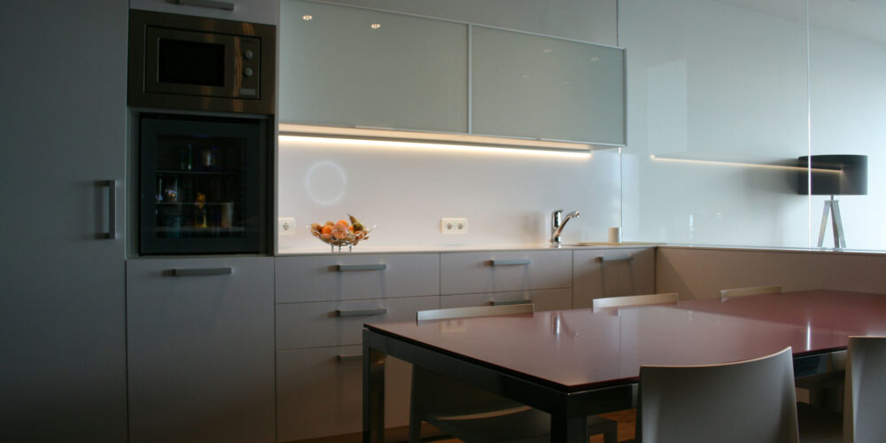 Interior de cocina moderna que presenta gabinetes blancos, diseño minimalista, microondas y horno empotrados, panel inferior de gabinete de vidrio negro, encimera brillante con un tazón de frutas y mesa de comedor con tapa de vidrio y sillas blancas, realzada por una suave iluminación superior y una pared espejada que aporta una sensación de amplitud.