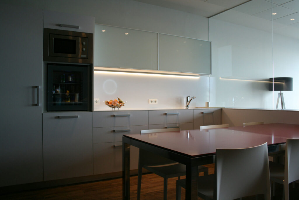 Cocina Moderna Minimalista con Elegantes Gabinetes Blancos y Área de Comedor Sofisticada Interior de cocina moderna que presenta gabinetes blancos, diseño minimalista, microondas y horno empotrados, panel inferior de gabinete de vidrio negro, encimera brillante con un tazón de frutas y mesa de comedor con tapa de vidrio y sillas blancas, realzada por una suave iluminación superior y una pared espejada que aporta una sensación de amplitud.