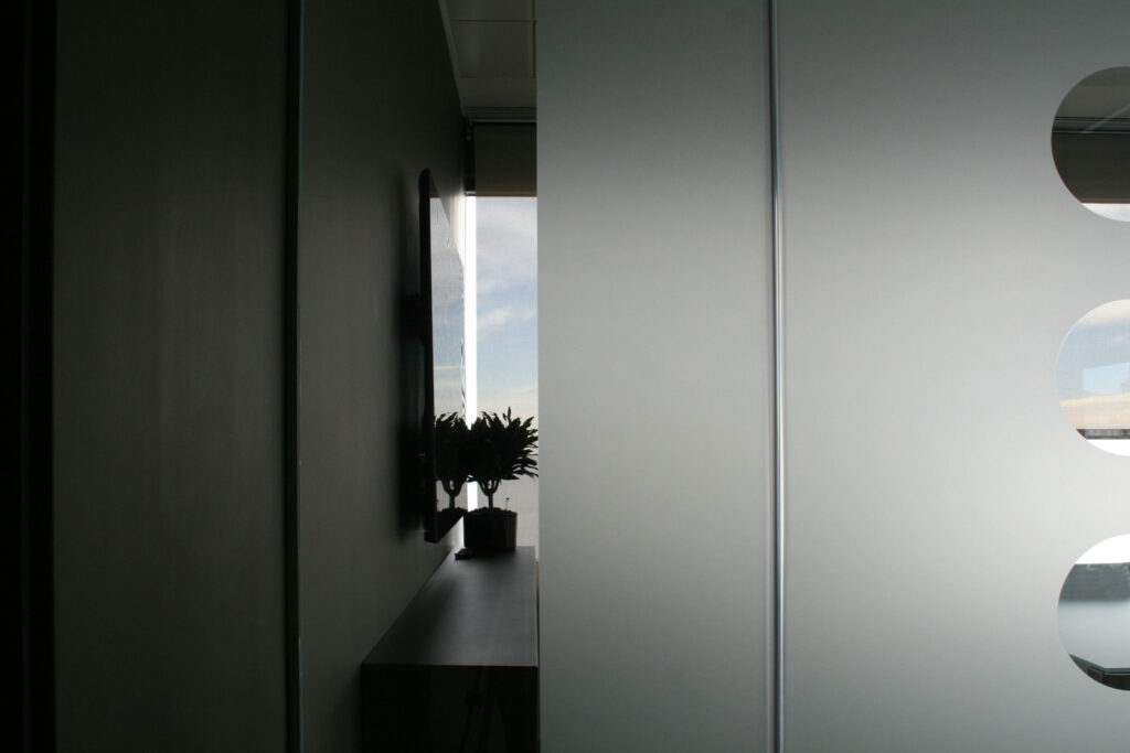 Diseño de Pasillo Moderno con Luz Natural y Vegetación Un sereno pasillo moderno con una pared izquierda adornada con una pequeña planta en una estantería contemporánea, y un lado derecho con una pared translúcida que presenta recortes circulares. La luz del sol entra a raudales a través de una ventana al final, realzando la atmósfera tranquila.