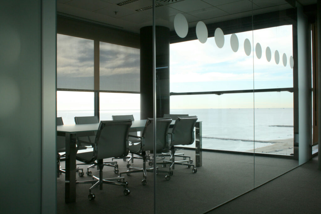 Sala de Conferencias Moderna con Vista al Mar y Asientos Ergonómicos Una sala de conferencias espaciosa con una mesa grande y sillas ergonómicas, diseñada con una decoración mínima y amplias ventanas que ofrecen vistas a un mar tranquilo. La luz natural se filtra a través de recortes circulares decorativos en las paredes de vidrio, creando una atmósfera abierta y serena para reuniones y discusiones.
