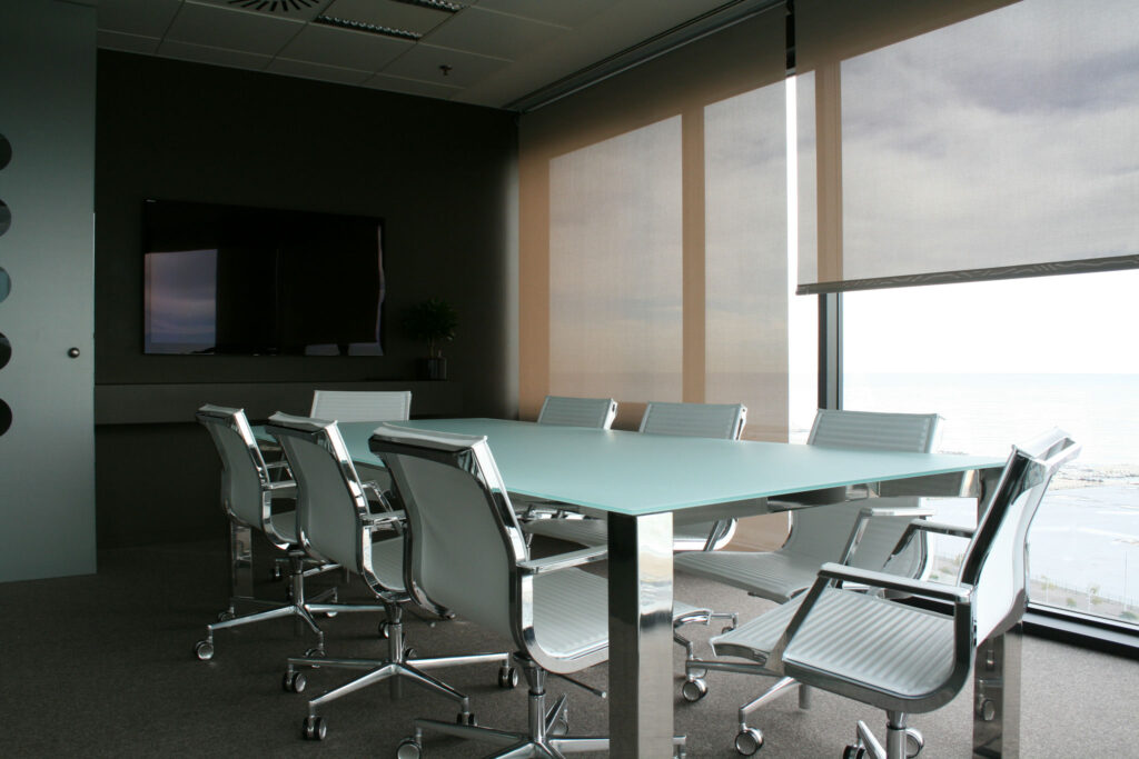 Sala de Conferencias Moderna con Mesa de Vidrio y Sillas Ergonómicas Una elegante sala de conferencias moderna que cuenta con una mesa de vidrio rodeada de sillas de oficina ergonómicas plateadas, bien iluminada con grandes ventanas y una decoración minimalista que incluye una pequeña planta.