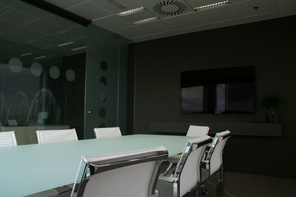 Sala de Conferencias Moderna con Mesa de Cristal y Diseño Minimalista Una elegante sala de conferencias moderna con una mesa de cristal rodeada de sillas blancas, una pared oscura y una decoración mínima que incluye una pequeña planta en maceta. La sala está bien iluminada con luz natural de una pared de cristal, creando una atmósfera profesional para reuniones y presentaciones.