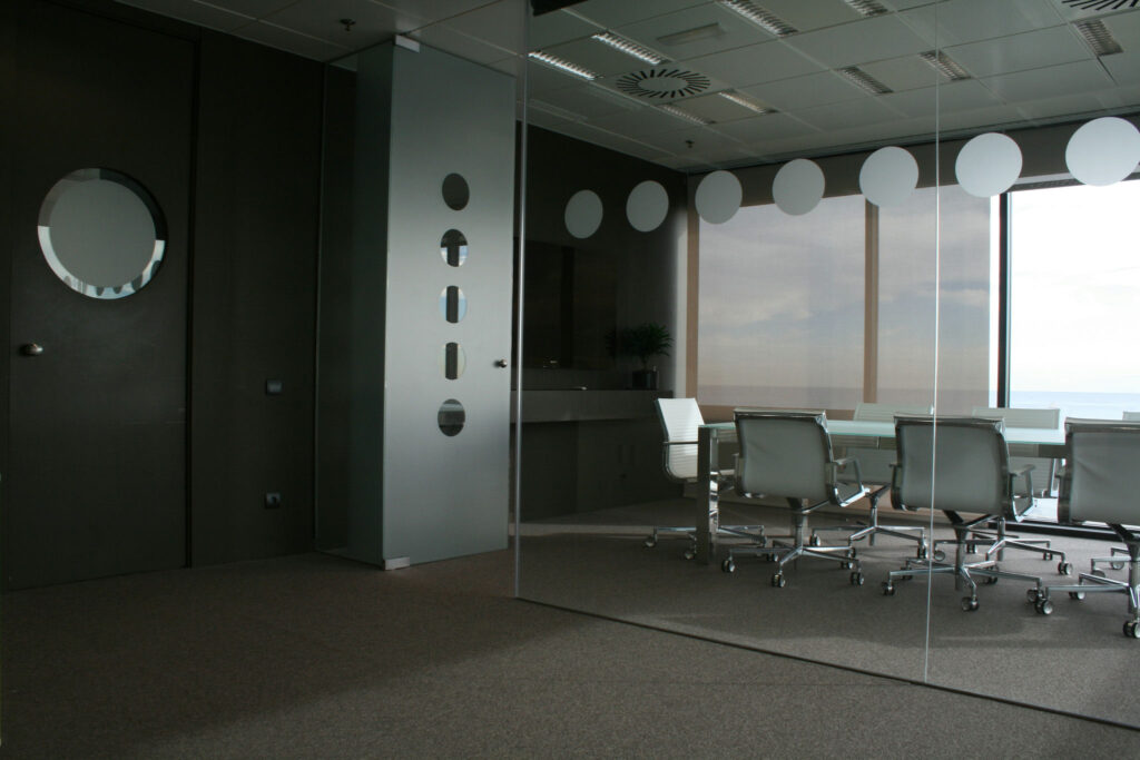 Sala de Conferencias Moderna con Paredes de Vidrio y Asientos Ergonómicos Una sala de conferencias moderna y elegante con paredes de vidrio, una gran mesa de conferencias rodeada de sillas ergonómicas, paneles de vidrio esmerilado con recortes circulares y grandes ventanas que ofrecen vistas al exterior. La sala tiene un cálido piso de alfombra y un esquema de color oscuro y profesional.