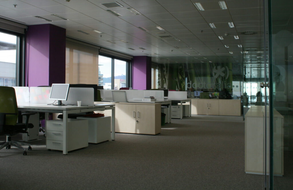 Diseño de Interiores de Oficina Moderna con Muebles Ergonómicos Un interior de oficina elegante que exhibe escritorios modernos completos con monitores de computadora, sillas ergonómicas y modernos armarios de almacenamiento blancos. Las grandes ventanas iluminan la habitación con luz natural, atrayendo la atención hacia la pared de acento púrpura. El espacio de trabajo está ordenado y organizado, con muebles de oficina adicionales visibles en el fondo, creando un entorno de trabajo profesional y acogedor.