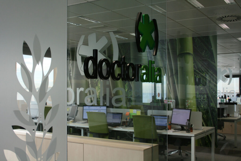 Entorno de Oficina Moderna con Logo de Doctoralia y Mural de Temática Natural Espacio de oficina brillante y contemporáneo que muestra el logo de Doctoralia en verde y negro, con paredes de vidrio, escritorios blancos limpios, mural vibrante de bambú y sillas verdes, enfatizando una atmósfera profesional pero conectada a la naturaleza para la tecnología de la salud.