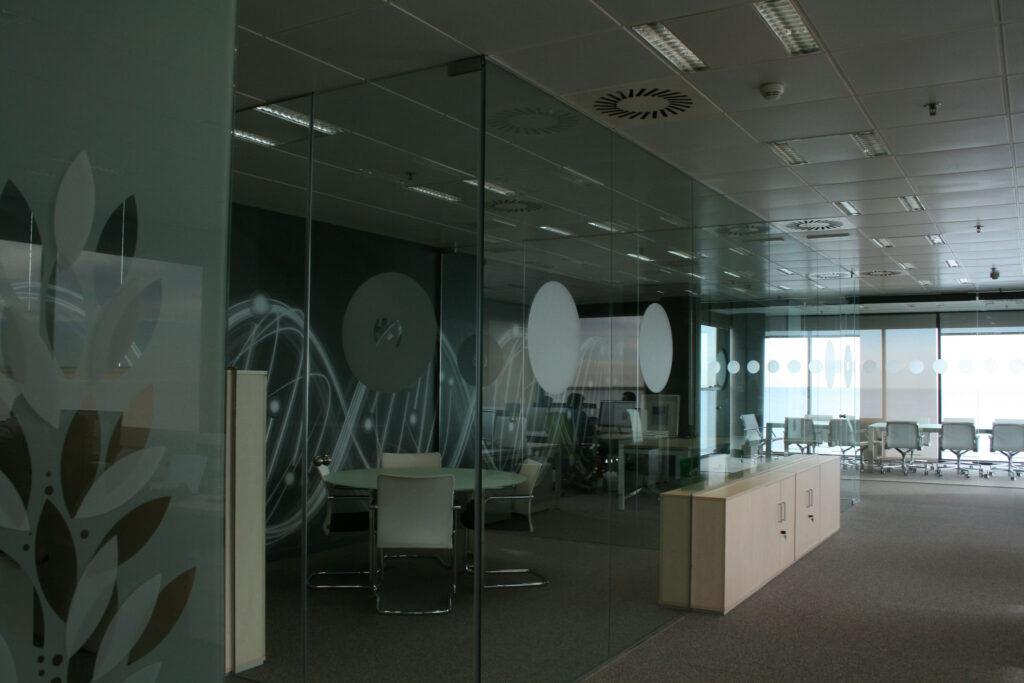 Interior de Oficina Moderna con Tabiques de Cristal y Diseño Minimalista Vista interior de una oficina moderna con tabiques de cristal, muebles de colores claros y motivos decorativos de hojas. Se puede ver una sala de reuniones espaciosa con sillas blancas y una mesa redonda, iluminada por la luz natural que entra a través de grandes ventanales.