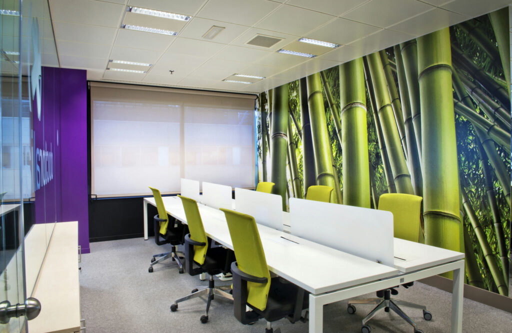 Sala de conferencias moderna con mural de bambú Una sala de conferencias moderna con un vibrante mural de bambú en una pared, equipada con elegantes escritorios blancos dispuestos colaborativamente y sillas ergonómicas verdes brillantes. La luz natural entra a raudales a través de grandes ventanas, creando una atmósfera acogedora.