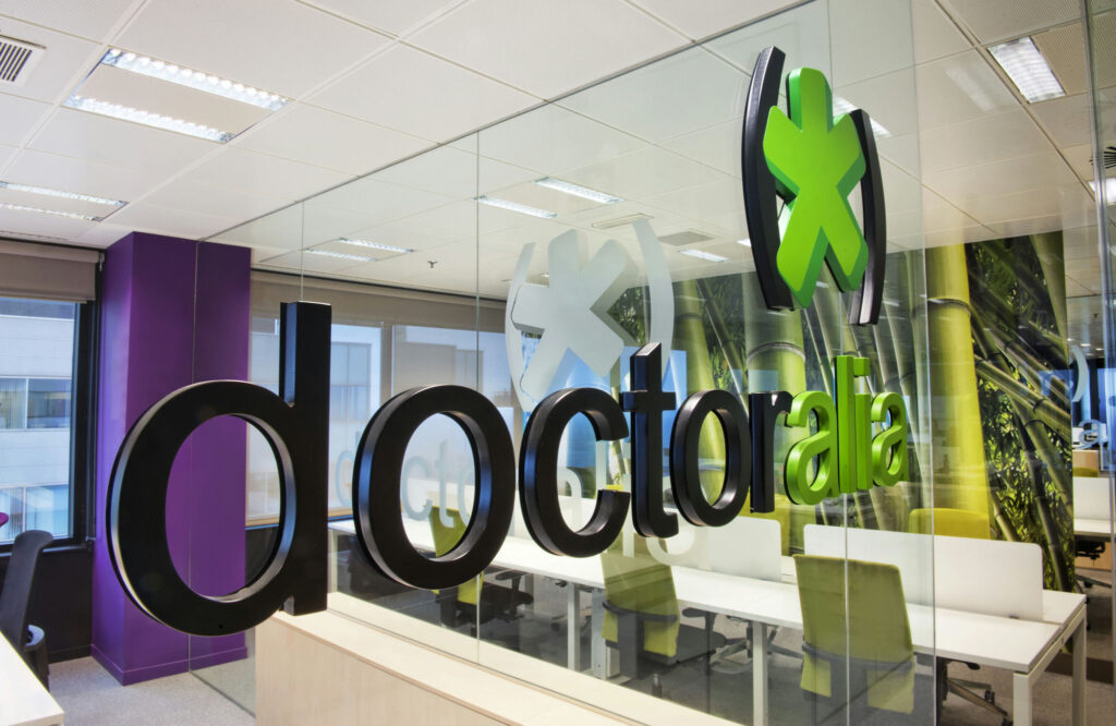 Espacio de oficina moderno de Doctoralia con diseño colaborativo Un interior de oficina moderno de Doctoralia con una pared de cristal, que muestra escritorios blancos, sillas verdes vibrantes y gráficos de bambú relajantes.