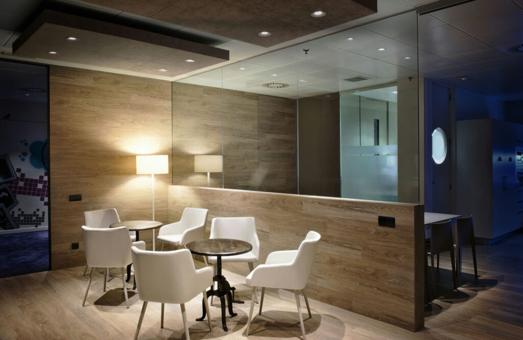Área de Asientos Moderna y Acogedora en Oficina con Sillas Blancas Elegantes Un interior de oficina moderno que muestra un área de asientos acogedora con cuatro elegantes sillas blancas alrededor de una mesa circular, rodeada de cálidas paredes de paneles de madera. Una partición de vidrio aporta un toque contemporáneo, mientras que la iluminación suave y una lámpara de pie crean una atmósfera acogedora.