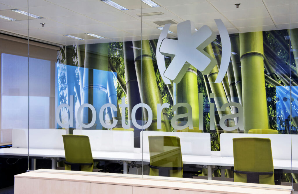 Oficinas Doctoralia: Un espacio de trabajo de oficina contemporáneo que presenta una pared de vidrio con la palabra "Doctoralia". Detrás de ella, un vibrante mural de tallos de bambú contra un cielo azul añade un toque orgánico al diseño interior moderno. El espacio de trabajo incluye elegantes escritorios blancos y cómodas sillas verdes, todo iluminado por una suave luz ambiental. Este entorno fomenta la colaboración y el profesionalismo, ideal para el trabajo en equipo.
