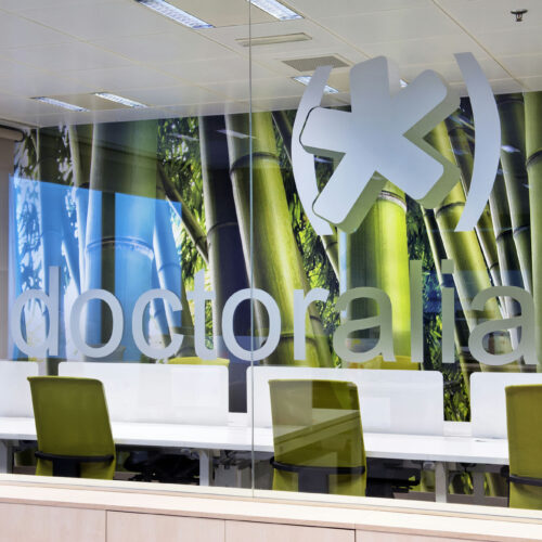 Oficinas Doctoralia: Un espacio de trabajo de oficina contemporáneo que presenta una pared de vidrio con la palabra "Doctoralia". Detrás de ella, un vibrante mural de tallos de bambú contra un cielo azul añade un toque orgánico al diseño interior moderno. El espacio de trabajo incluye elegantes escritorios blancos y cómodas sillas verdes, todo iluminado por una suave luz ambiental. Este entorno fomenta la colaboración y el profesionalismo, ideal para el trabajo en equipo.