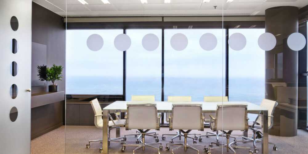 Una amplia sala de conferencias moderna con una gran mesa blanca rodeada de elegantes sillas plateadas, paredes de vidrio con diseños circulares esmerilados y una vista panorámica del océano. La luz natural inunda la sala, realzando la atmósfera acogedora, con una planta verde en una repisa que añade un toque de naturaleza.
