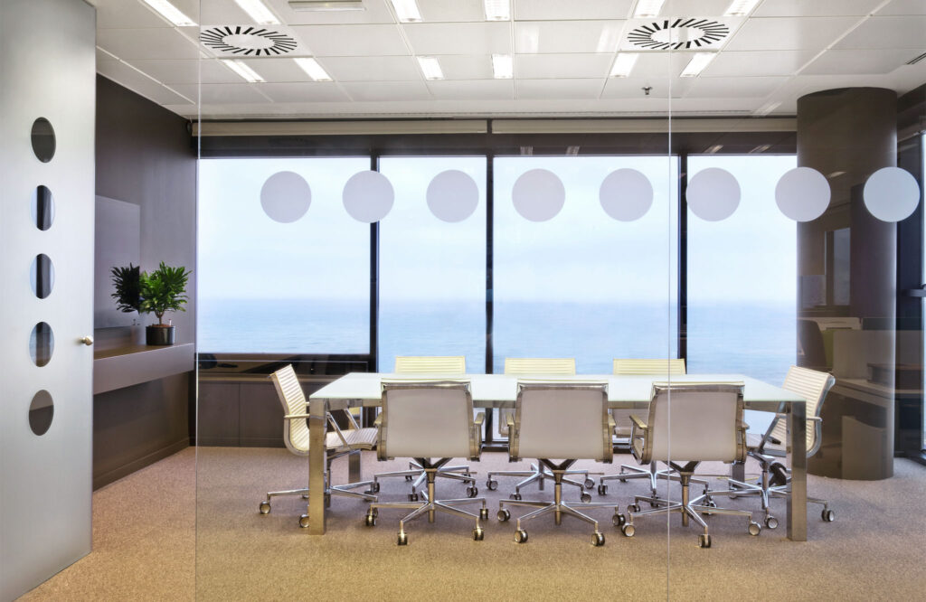 Sala de Conferencias Moderna con Vista Escénica al Océano Una sala de conferencias contemporánea con una pared de vidrio adornada con diseños circulares, una gran mesa rectangular rodeada de sillas de oficina ergonómicas, y amplias ventanas que muestran una hermosa vista al océano. La sala está iluminada con luz superior e incluye una planta decorativa para añadir un toque de verde.