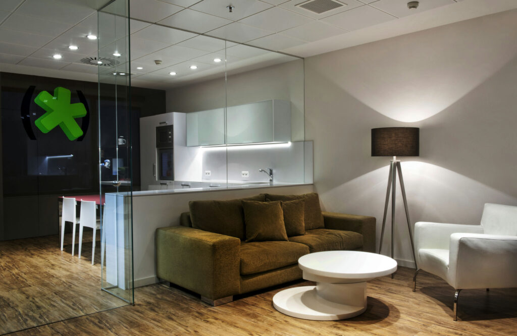 Espacio de Vida Moderno y Estiloso con Enclavamiento de Vidrio y Diseño Minimalista Una sala de estar contemporánea que presenta un espacio cerrado con vidrio y elementos de diseño minimalista. Un sofá marrón y una silla blanca están ubicados cerca de una mesa de café redonda, complementados por una iluminación suave. En el fondo, hay un área de comedor con sillas blancas adyacente a una cocina elegante que cuenta con gabinetes y electrodomésticos blancos, todo ello sobre un cálido suelo de madera.
