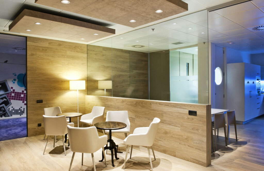 Área de Reunión de Oficina Moderna con Pared de Vidrio y Acabados en Madera Cálida Un espacio de oficina moderno que muestra una acogedora área de reunión con una mesa redonda y cuatro sillas blancas, iluminada por una lámpara de pie. El espacio está separado por una gran pared de vidrio, con tonos cálidos de madera en las paredes y una iluminación superior que crea una atmósfera acogedora. Se puede ver un vistazo de muebles de oficina y decoraciones adicionales en el fondo.