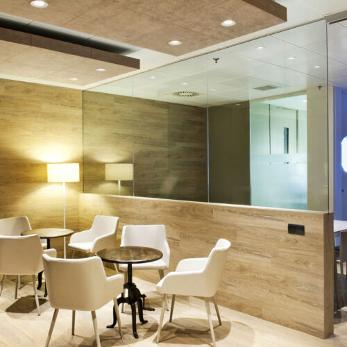 Un espacio de oficina moderno que muestra una acogedora área de reunión con una mesa redonda y cuatro sillas blancas, iluminada por una lámpara de pie. El espacio está separado por una gran pared de vidrio, con tonos cálidos de madera en las paredes y una iluminación superior que crea una atmósfera acogedora. Se puede ver un vistazo de muebles de oficina y decoraciones adicionales en el fondo.